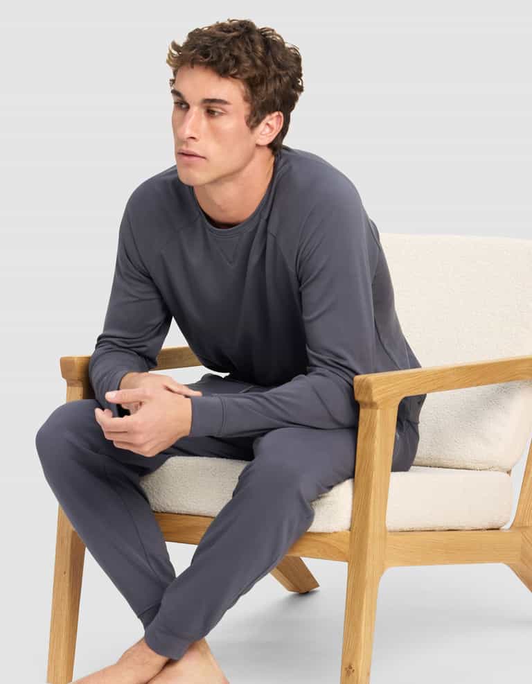 A young man sits barefoot on a cushioned wooden chair, wearing Cozy Earth's Men's Brushed Bamboo Jogger Pant in gray with a matching long-sleeve top. He looks forward with clasped hands against a plain, light gray background.|Color:Graphite