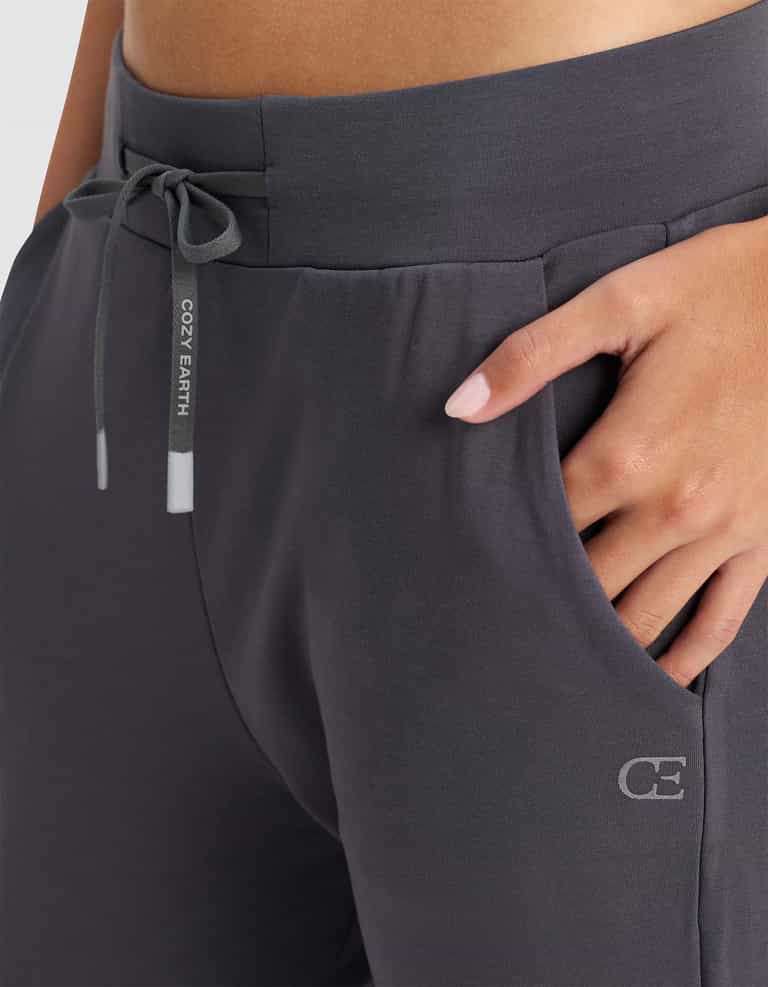 A woman wears Cozy Earth's Women's Brushed Bamboo Jogger Pant in dark gray, featuring "COZY EARTH" on the drawstring and a "CE" logo near the pocket, with one hand casually placed in the pocket.|Color:Graphite