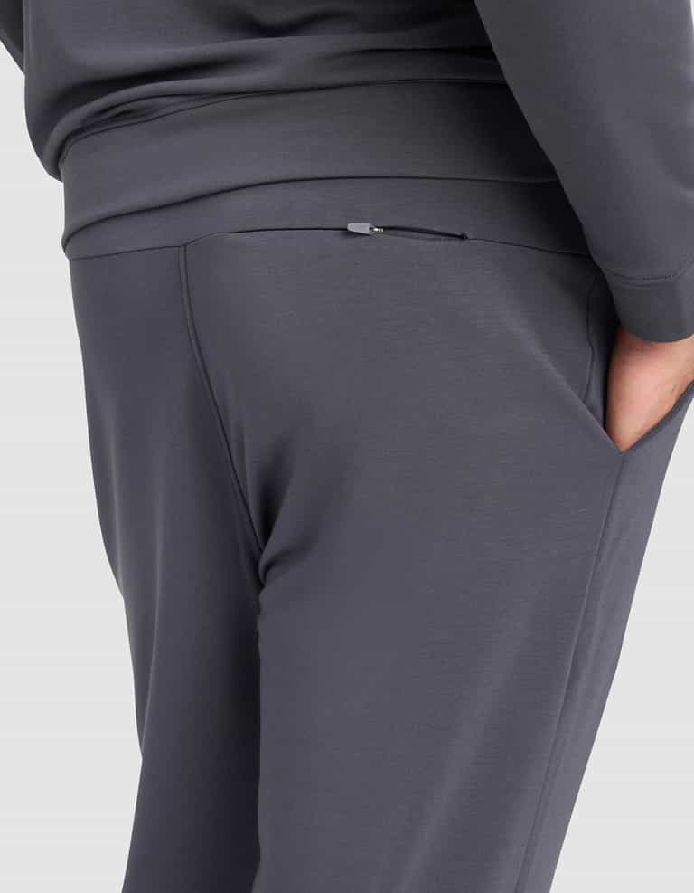 A person wearing Cozy Earth Men's Brushed Bamboo Jogger Pant in dark gray, with one hand in the pocket, showcases a close-up of the waist and upper legs plus a visible back zipper detail.|Color:Graphite