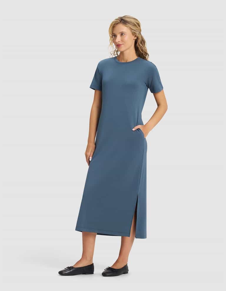 A woman wears the Cozy Earth Women's Brushed Bamboo Midi Dress—short-sleeve, ankle-length, blue with a side slit and pockets—standing against a plain light background and smiling slightly. She pairs it with black flats.|Color:Dawn