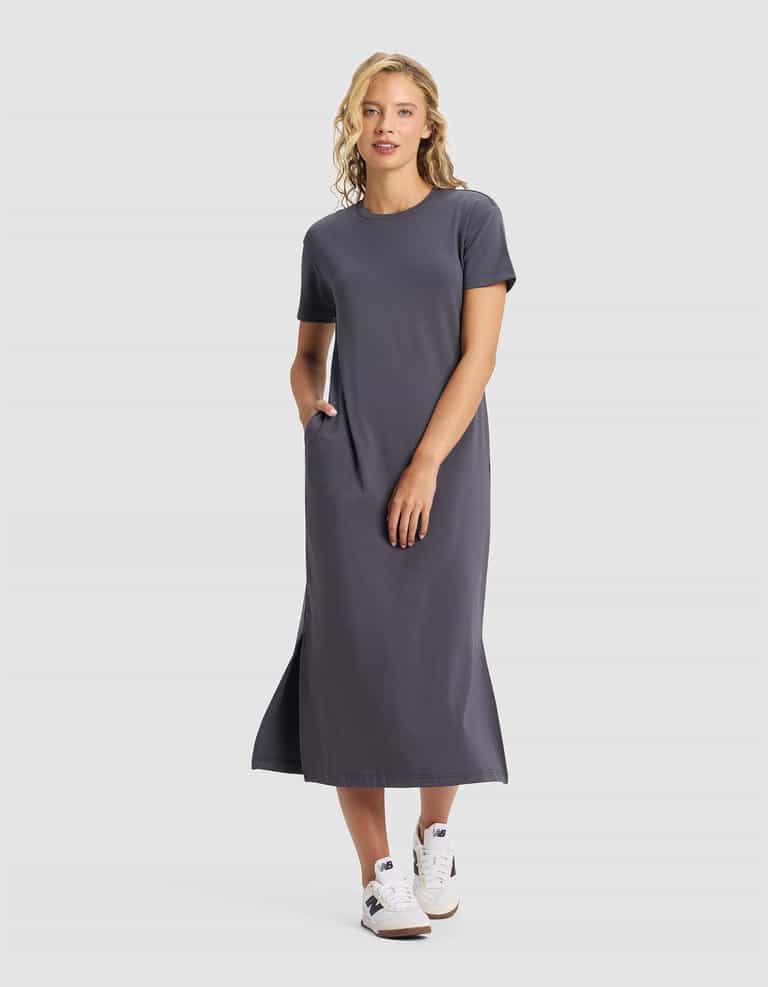 A woman smiles against a plain background, wearing the Cozy Earth Women's Brushed Bamboo Midi Dress in gray with short sleeves, side pockets, and white sneakers. She has one hand in her pocket.|Color:Graphite