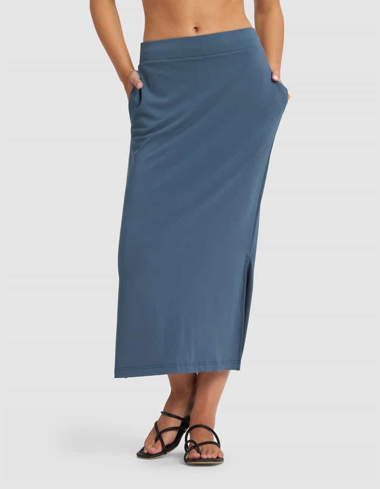 A person wears the Cozy Earth Women's Brushed Bamboo Midi Skirt—a high-waisted, ankle-length blue skirt with side pockets and a side slit—paired with black strappy sandals against a plain white background.|Color:Dawn