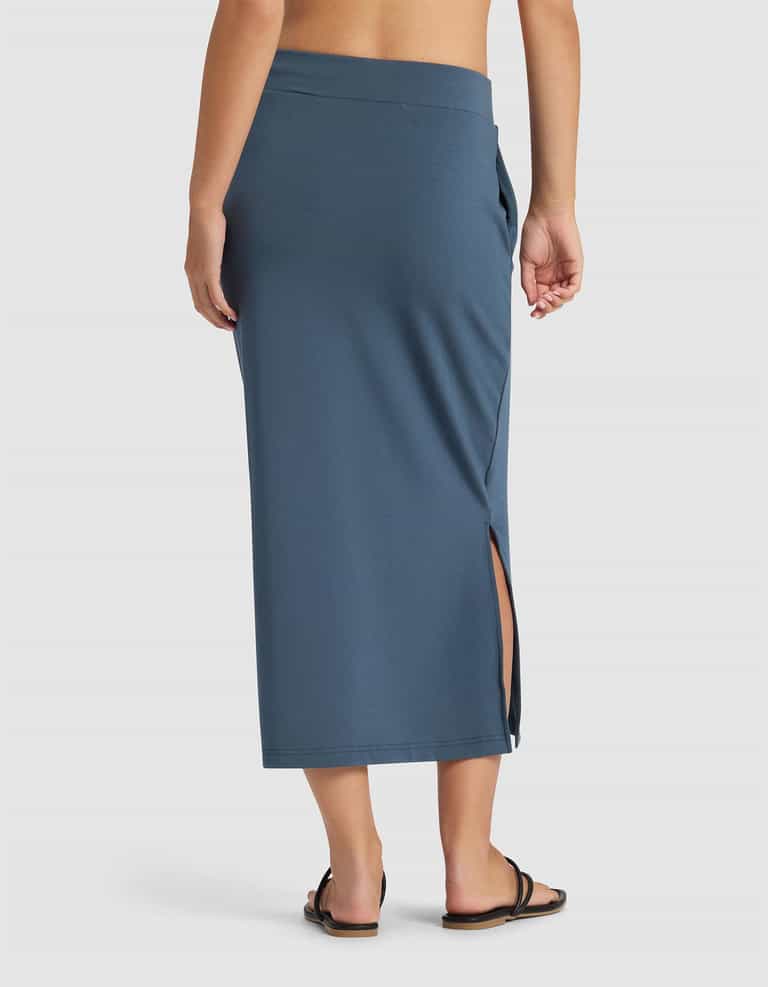 A woman is shown from the back wearing Cozy Earth’s Women’s Brushed Bamboo Midi Skirt in blue with a side slit, paired with black sandals, set against a plain white background.|Color:Dawn