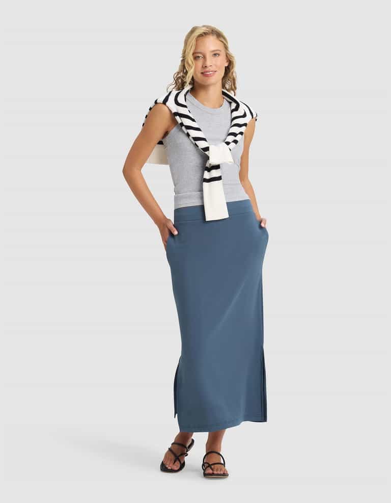 A woman smiles with hands in her pockets, wearing a grey sleeveless top, black sandals, and Cozy Earth's Women's Brushed Bamboo Midi Skirt in blue. A white sweater with black stripes is draped over her shoulders.|Color:Dawn