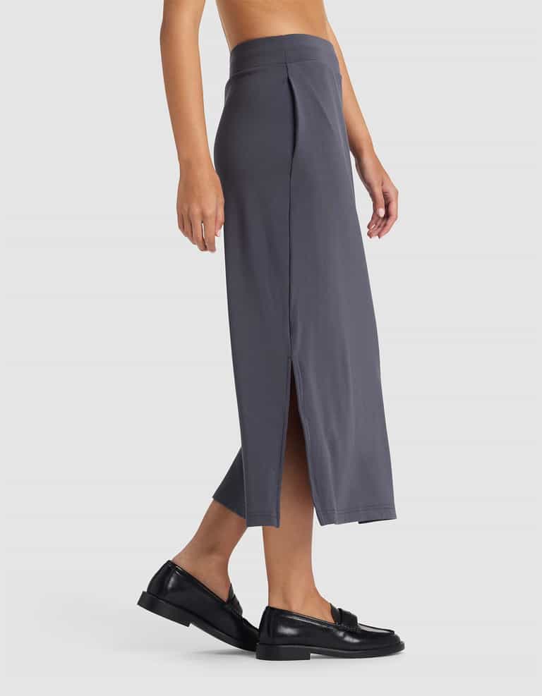 A person is seen from the side wearing Cozy Earth’s Women’s Brushed Bamboo Midi Skirt in gray, featuring a high waist and side slit, paired with black loafers against a light gray background.|Color:Graphite