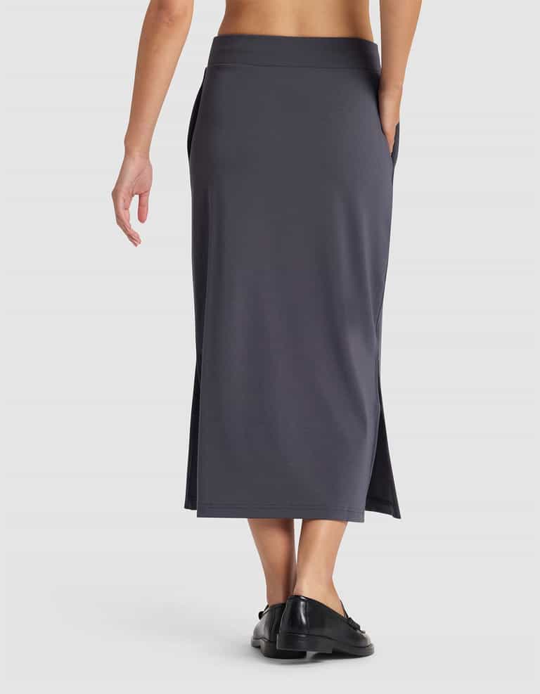 A person is shown from the back wearing the Cozy Earth Women's Brushed Bamboo Midi Skirt in dark gray with side slits and black shoes, set against a plain white background.|Color:Graphite
