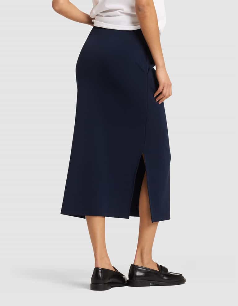A woman is shown from the back wearing the Cozy Earth Women's Brushed Bamboo Midi Skirt in navy blue with a side slit, paired with a white top and black loafers, against a plain light gray background. |Color:Navy