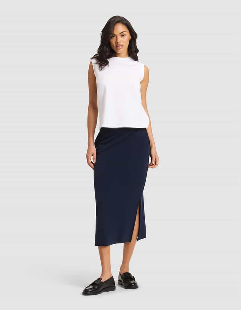 A woman stands against a plain background wearing a sleeveless white top, black loafers, and the Cozy Earth Women's Brushed Bamboo Midi Skirt in navy blue with a side slit. |Color:Navy