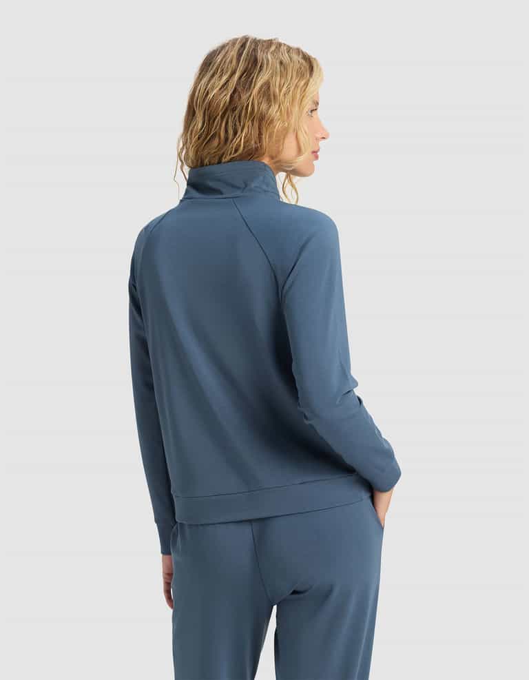 A woman with wavy blonde hair faces away, wearing the Cozy Earth Women's Brushed Bamboo Mock Neck and matching blue pants. Her hands are in her pockets against a plain light gray background.|Color:Dawn
