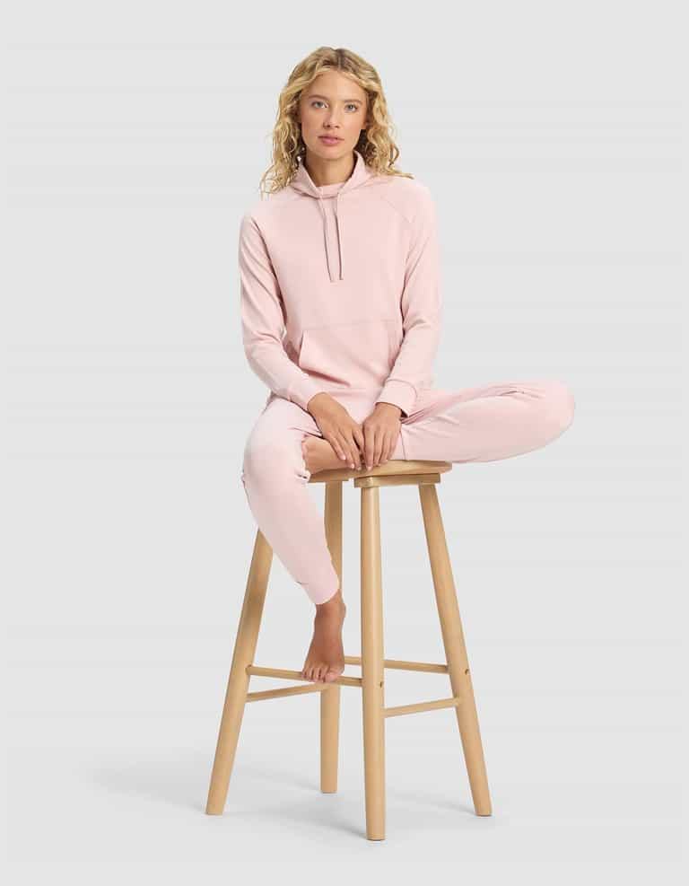 A woman wearing the Cozy Earth Women's Brushed Bamboo Mock Neck sits barefoot on a wooden stool, one leg bent and the other hanging down, against a plain light gray background.|Color:Dusty Rose