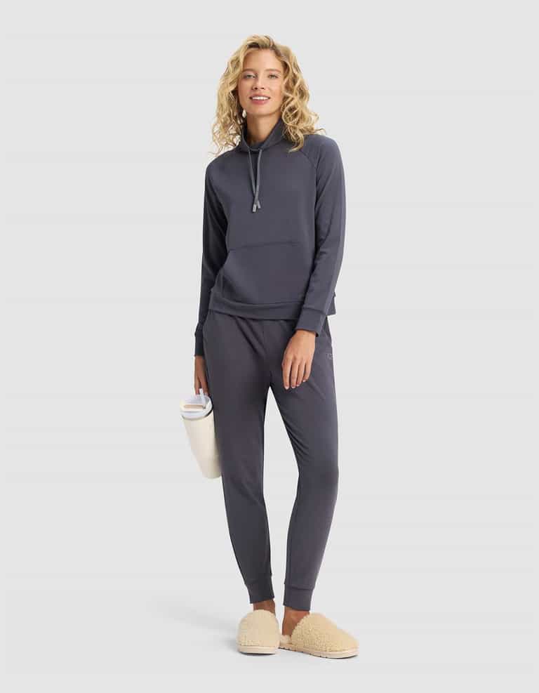 A smiling woman wears the Cozy Earth Women's Brushed Bamboo Mock Neck in dark gray with tan slippers, holding a white water bottle against a plain light background.|Color:Graphite