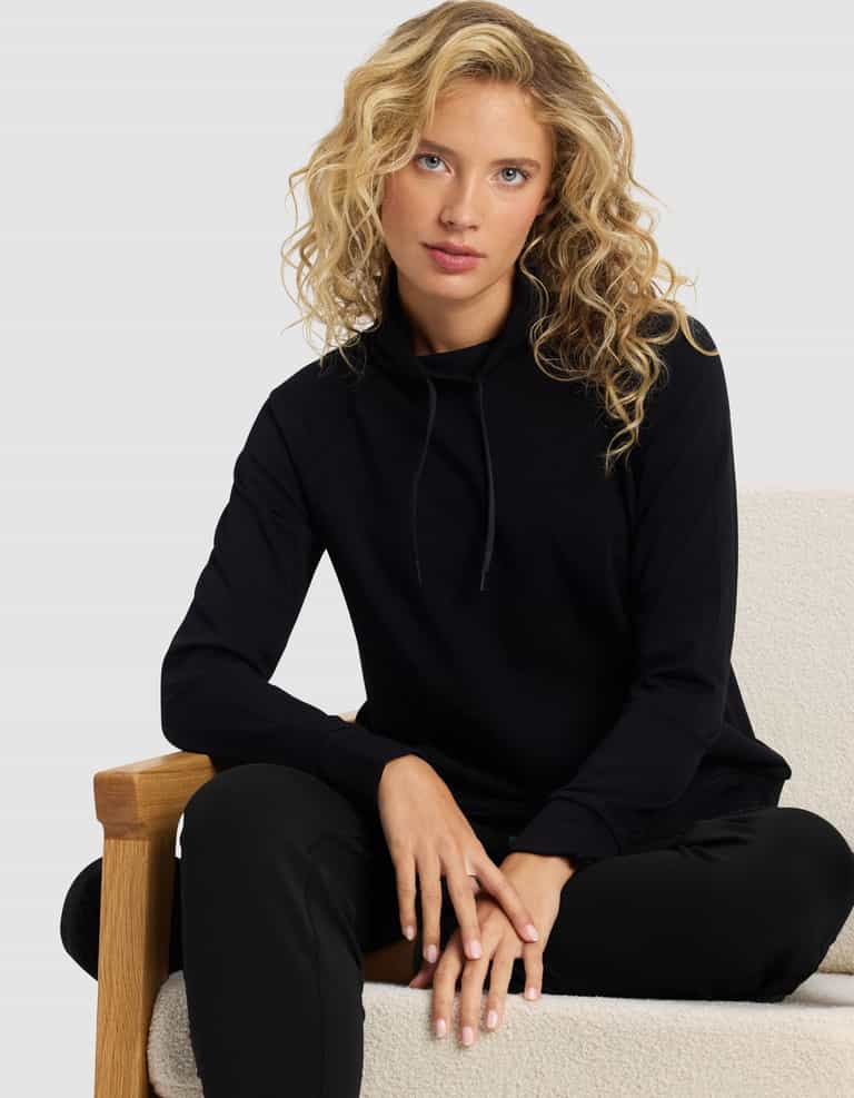 A woman with curly blonde hair wears the Cozy Earth Women's Brushed Bamboo Mock Neck & Jogger Set, sitting on a light cushioned chair with one arm resting on the wooden armrest.|Color:Black