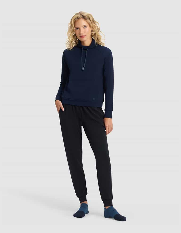 A woman with curly blonde hair wears the Cozy Earth Women's Brushed Bamboo Mock Neck, paired with black jogger pants and navy socks, standing against a plain light gray background with a relaxed posture and a slight smile.|Color:Navy
