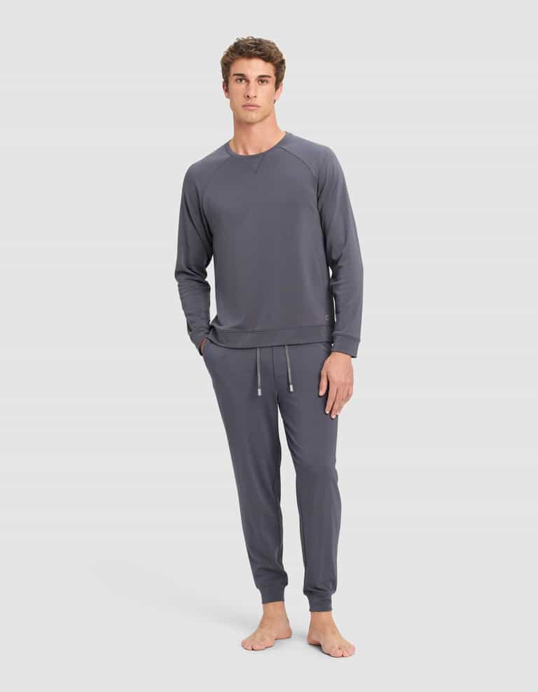 A man with short curly hair and a relaxed expression stands barefoot against a plain background, wearing the Cozy Earth Men's Brushed Bamboo Jogger Set in slate gray, featuring a long-sleeve top and drawstring jogger-style pants.|Color:Graphite