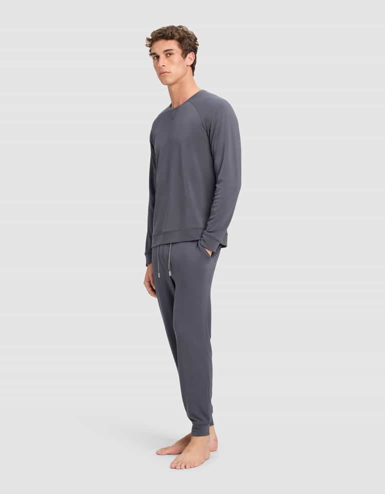 A man with short curly hair stands barefoot against a plain background, wearing Cozy Earth's Men's Brushed Bamboo Jogger Pant in dark gray with a relaxed-fit long-sleeve top. He has a neutral expression.|Color:Graphite