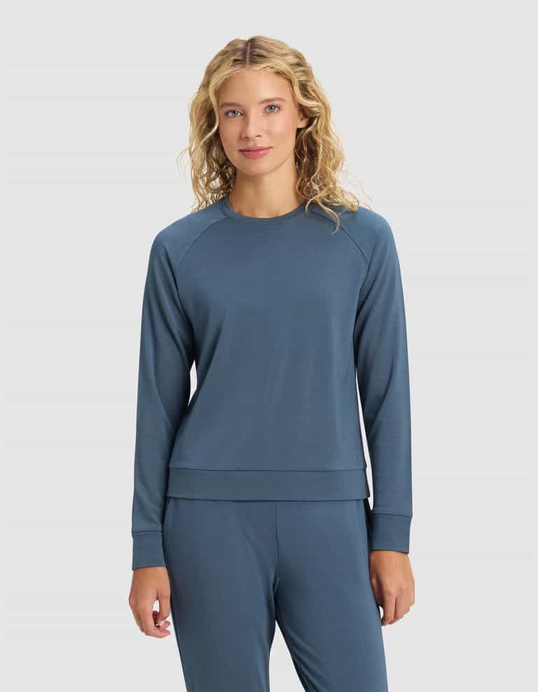 A woman with curly blonde hair faces forward against a plain light gray background, wearing the Cozy Earth Women's Brushed Bamboo Pullover Crew in blue, paired with matching pants.|Color:Dawn