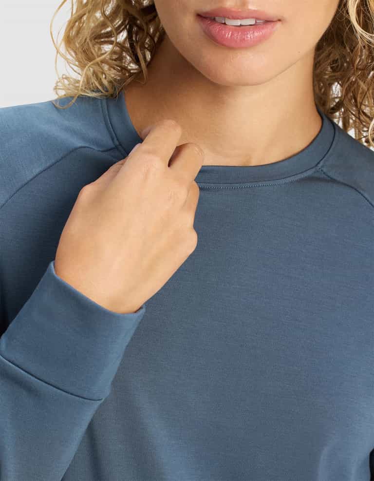A woman with curly blonde hair wears a blue Cozy Earth Women's Brushed Bamboo Pullover Crew, holding the collar with one hand; only her shoulders to lips are visible.|Color:Dawn