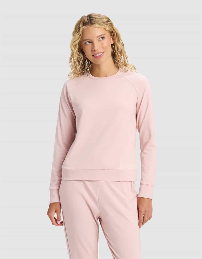 A woman with blonde, wavy hair wears the Cozy Earth Women's Brushed Bamboo Pullover Crew in light pink, paired with matching pants, standing against a plain light gray background and smiling softly while looking to the side.|Color:Dusty Rose