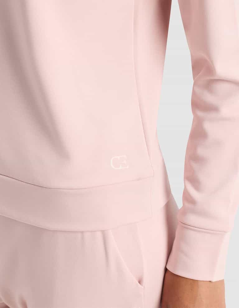 A woman wears the Cozy Earth Women's Brushed Bamboo Pullover Crew in light pink with matching pants, featuring a smooth fabric and a small white "CE" logo on the lower side of the top. One hand rests in her pocket.|Color:Dusty Rose