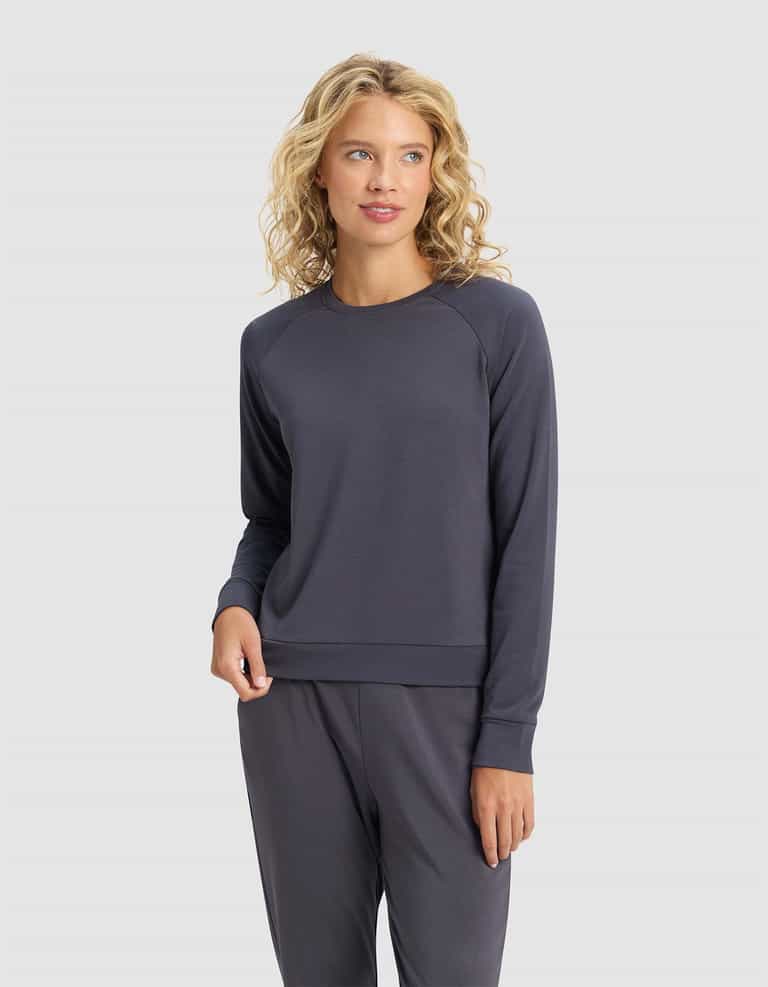 A woman with curly blonde hair wears the Cozy Earth Women's Brushed Bamboo Pullover Crew in dark gray, standing against a plain light gray background.|Color:Graphite