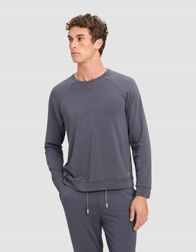 A man wears the Cozy Earth Men's Brushed Bamboo Pullover Crew in gray, paired with matching jogger pants, standing against a plain light background and looking slightly left with one hand in his pocket.|Color:Graphite