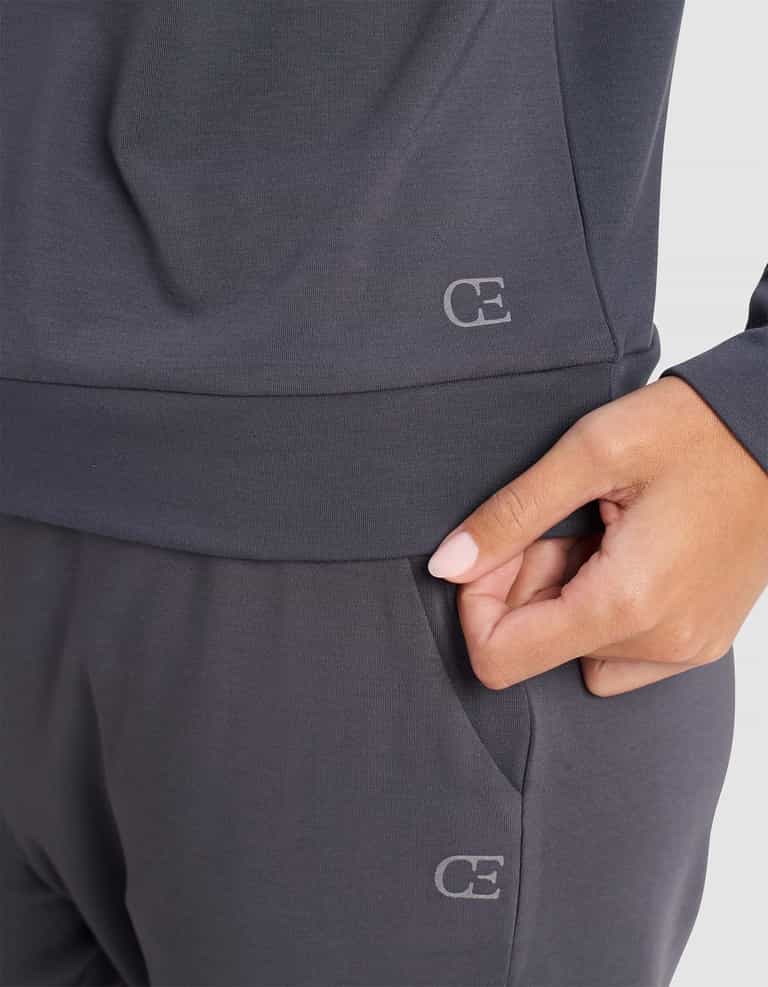 A person wears the Cozy Earth Women's Brushed Bamboo Pullover Crew in dark gray, with a subtle "CE" logo and their right hand in the pants pocket, paired with matching bottoms.|Color:Graphite