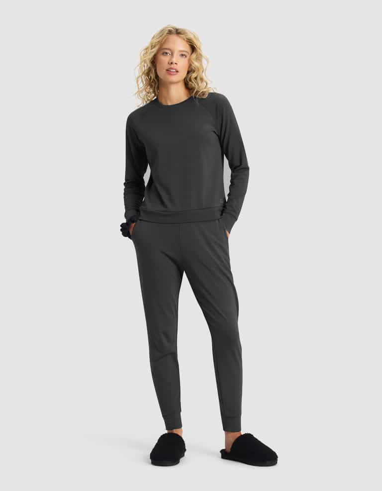 A woman with long, curly blond hair models the Cozy Earth Women's Brushed Bamboo Jogger Set in dark gray and black slippers, standing hands in pockets against a plain light gray background.|Color:Charcoal