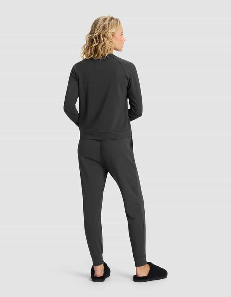 A woman with blonde, wavy hair faces away, wearing the Cozy Earth Women's Brushed Bamboo Jogger Set in dark gray with black slippers, against a plain light gray background.|Color:Charcoal