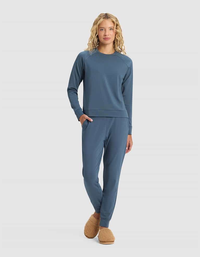 A woman with long curly blonde hair models the Cozy Earth Women's Brushed Bamboo Jogger Set in blue, standing with one hand in her pocket and wearing brown fuzzy slippers against a plain light background.|Color:Dawn