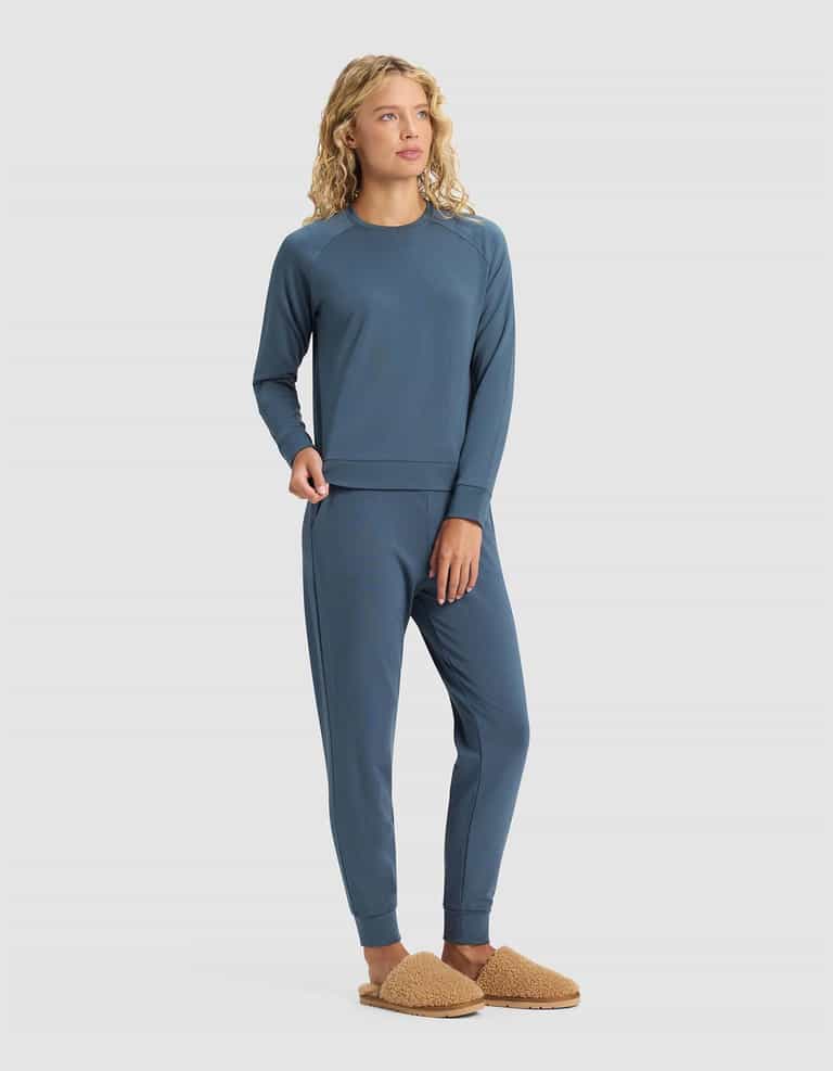 A woman with curly blonde hair wears the Cozy Earth Women's Brushed Bamboo Jogger Set in blue with tan fuzzy slippers, standing against a plain light background.|Color:Dawn