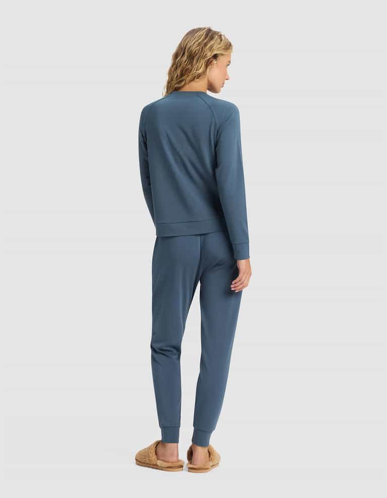 A person with wavy blonde hair stands facing away, wearing the Cozy Earth Women's Brushed Bamboo Jogger Set in blue and tan fuzzy slippers, set against a plain light background.|Color:Dawn