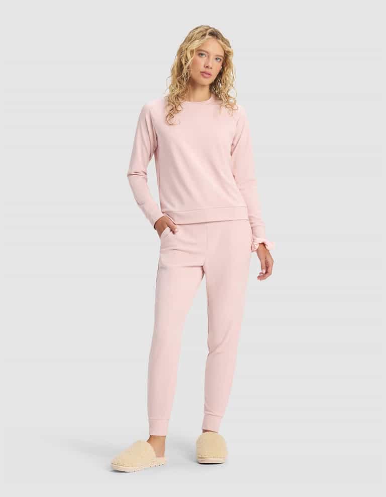 A woman with long, curly blonde hair wears the Cozy Earth Women's Brushed Bamboo Jogger Set in light pink with beige fuzzy slippers, standing against a plain light gray background.|Color:Dusty Rose