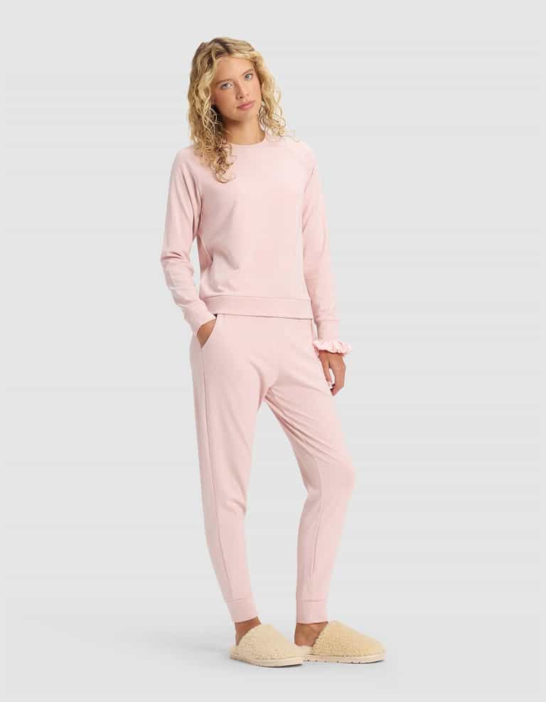 A young woman with curly blonde hair models the Cozy Earth Women's Brushed Bamboo Jogger Set in light pink, paired with beige fuzzy slippers, standing against a plain light gray background with one hand in her pocket.|Color:Dusty Rose