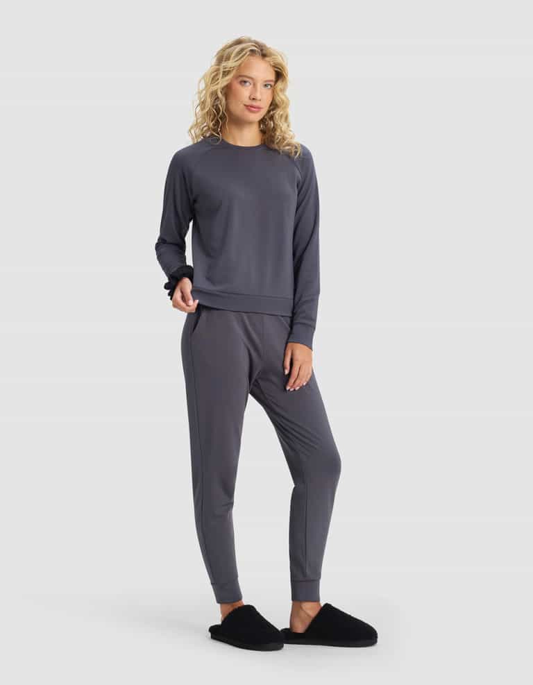 A woman with long curly blonde hair models the Cozy Earth Women's Brushed Bamboo Jogger Set in charcoal gray, paired with black slippers, standing against a plain light gray background.|Color:Graphite