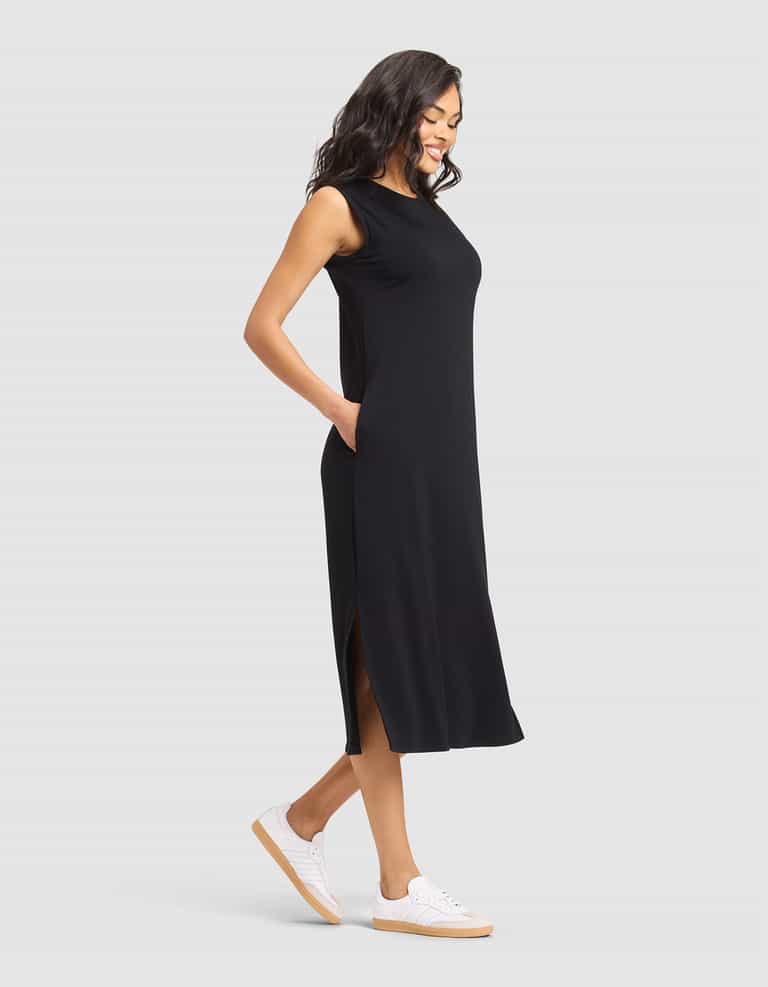 A woman smiles with her hands in her pockets, wearing the Cozy Earth Women's Brushed Bamboo Sleeveless Midi Dress and white sneakers, standing against a plain light gray background. |Color:Black