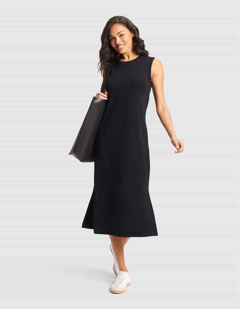 A woman smiles against a light gray background wearing Cozy Earth's Women's Brushed Bamboo Sleeveless Midi Dress in black, paired with white sneakers and a large black tote bag. |Color:Black