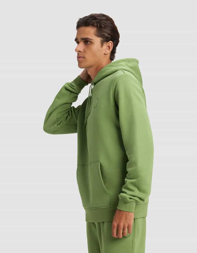 A young man in profile wears the Cozy Earth Men's Cityscape 2.0 Hoodie in light green, paired with matching joggers. His left hand touches the back of his neck against a plain light gray background.|Color:Clover