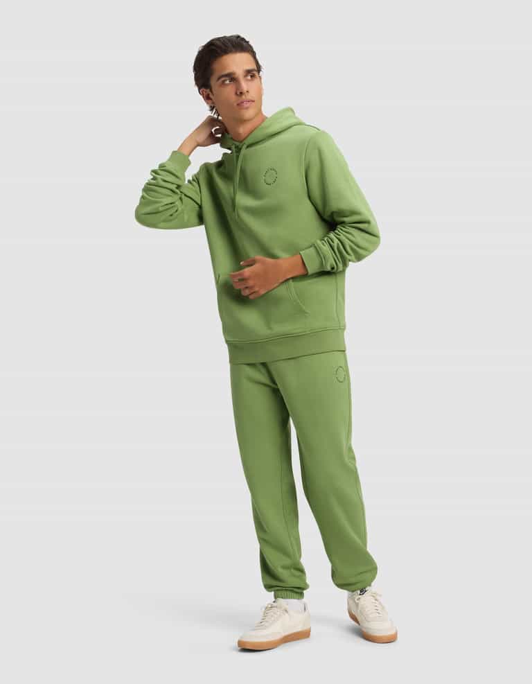 A young man in the Cozy Earth Men's Cityscape 2.0 Hoodie and matching sweatpants with white sneakers stands against a plain background, looking to the side with one hand touching his neck.|Color:Clover