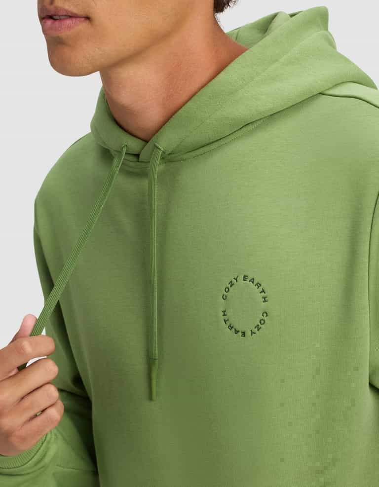 A man wears the Men's Cityscape 2.0 Hoodie by Cozy Earth in light green, featuring a small circular logo on the chest. He holds a hoodie drawstring, with only his lower face visible.|Color:Clover