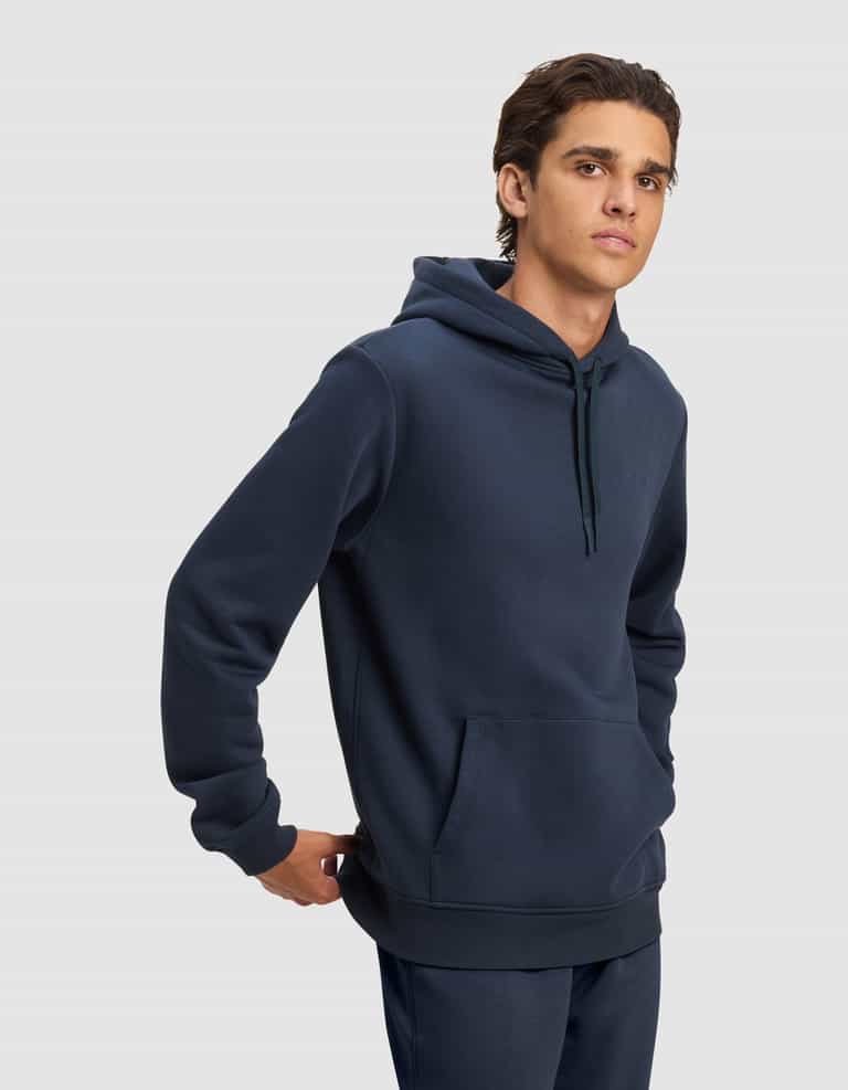 A young man models the Cozy Earth Men's Cityscape 2.0 Hoodie in dark blue with matching pants, standing with one hand behind his back and gazing to the side against a plain light gray background.|Color:Eclipse