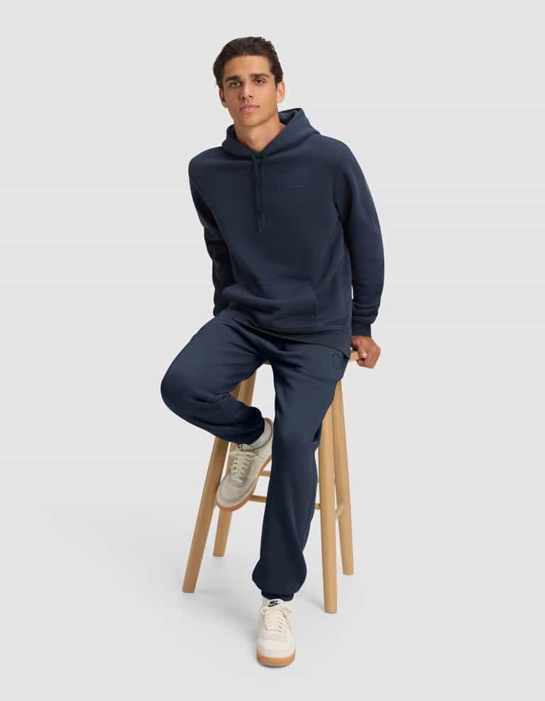 A man wears the Cozy Earth Men's Cityscape 2.0 Hoodie and matching sweatpants, sitting on a wooden stool against a plain light gray background, with one foot on the stool’s lower rung and white sneakers visible.|Color:Eclipse