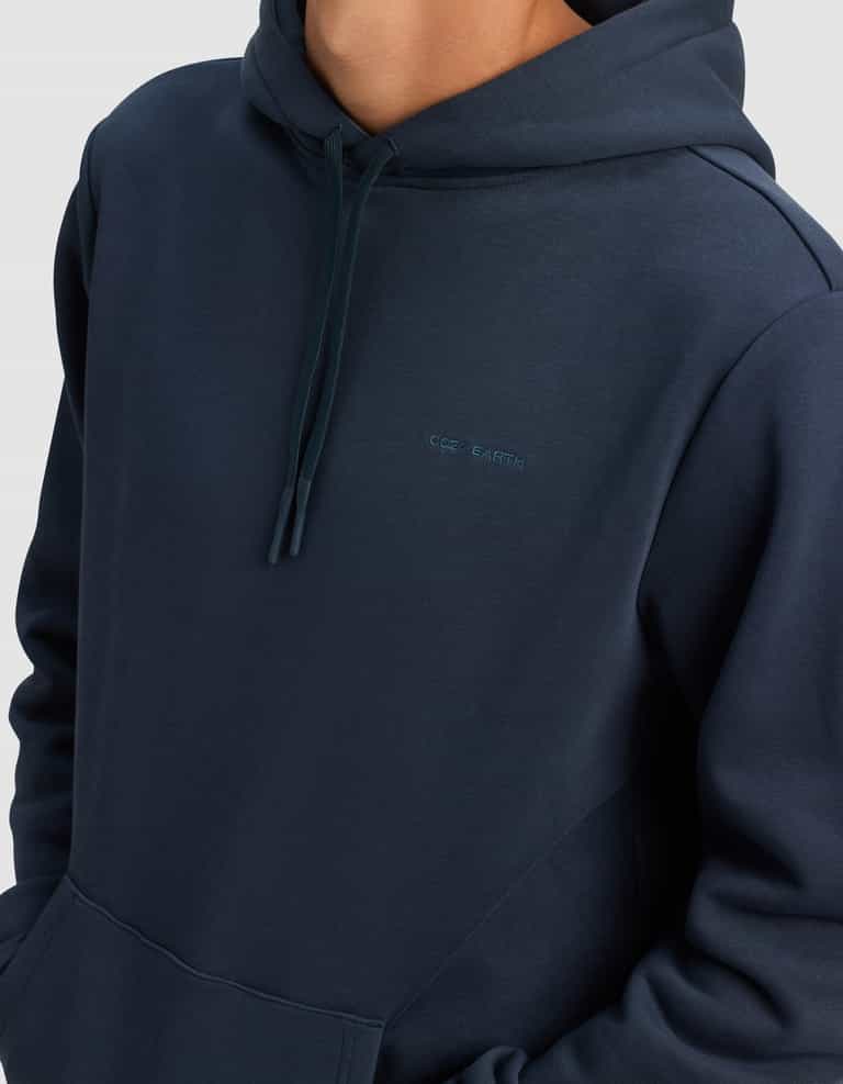 A person wears the Men's Cityscape 2.0 Hoodie by Cozy Earth—a dark blue hoodie with a front pocket, drawstrings, and "COS EARTH" embroidered on the chest. The person’s face is not shown.|Color:Eclipse