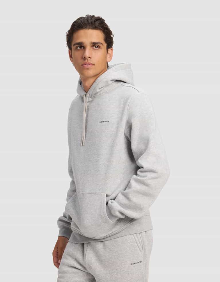 A young man models the Cozy Earth Men's Cityscape 2.0 Hoodie in light gray, with matching sweatpants, standing against a plain background and gazing slightly to the side with one hand in his pocket.|Color:Heather Grey