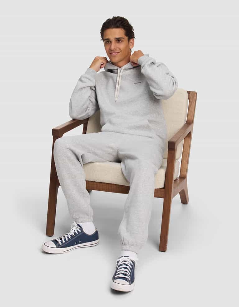 A young man relaxes on a cushioned chair, smiling slightly as he holds the drawstrings of his Cozy Earth Men's Cityscape 2.0 Hoodie in light gray, paired with sweatpants and dark blue sneakers.|Color:Heather Grey