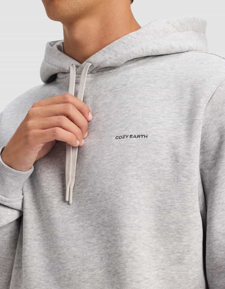 A person wearing the Cozy Earth Men's Cityscape 2.0 Hoodie in light gray holds the drawstrings near their neck against a plain, light-colored background. The "Cozy Earth" logo is visible on the chest.|Color:Heather Grey