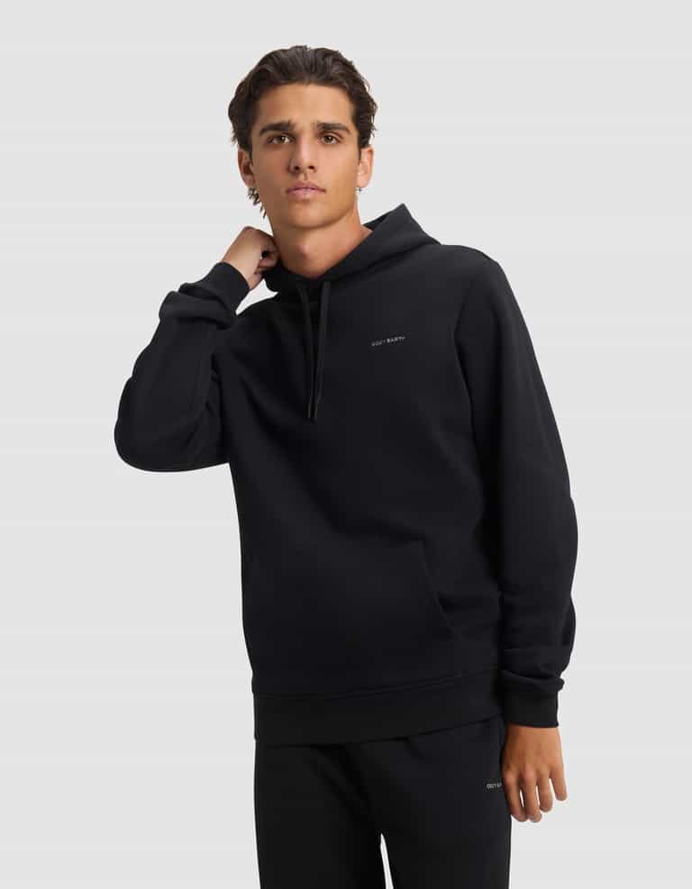 A young man models the Cozy Earth Men's Cityscape 2.0 Hoodie in black, paired with matching pants, standing against a light background with one hand on his neck and a neutral expression.|Color:Jet Black