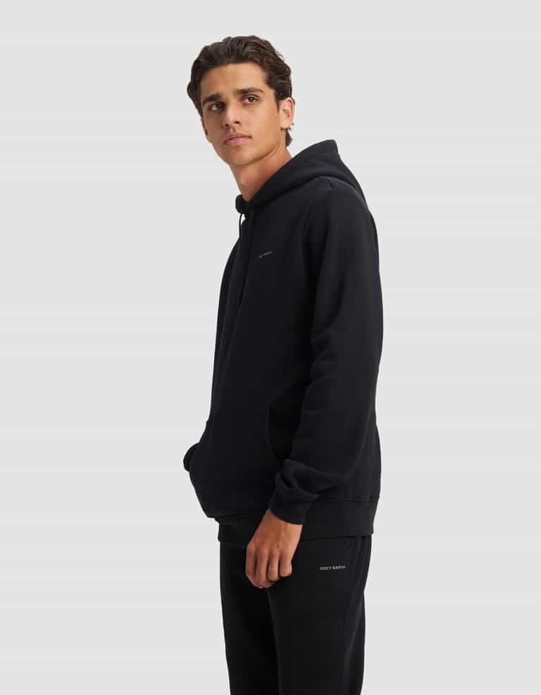 A young man with short brown hair wears the Cozy Earth Men's Cityscape 2.0 Hoodie and black pants, standing against a plain light gray background and looking slightly to his left with one hand in his hoodie pocket.|Color:Jet Black