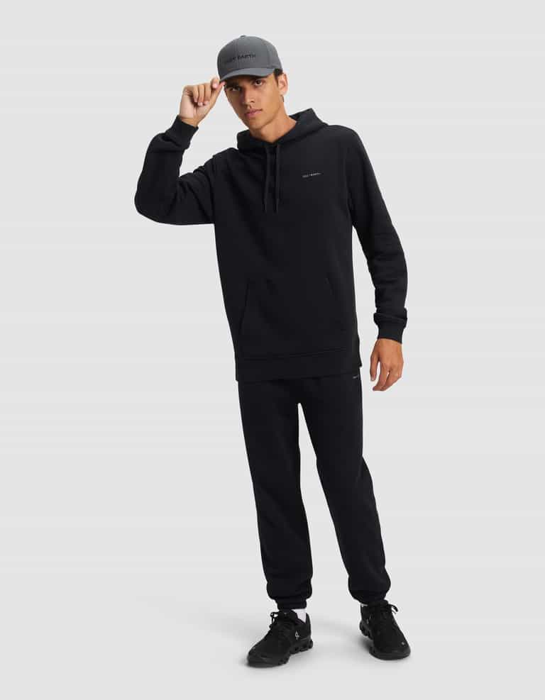 A person wearing the Cozy Earth Men's Cityscape 2.0 Hoodie stands against a plain background, dressed in black joggers, black sneakers, and a gray cap, holding the brim of the cap with one hand.|Color:Jet Black