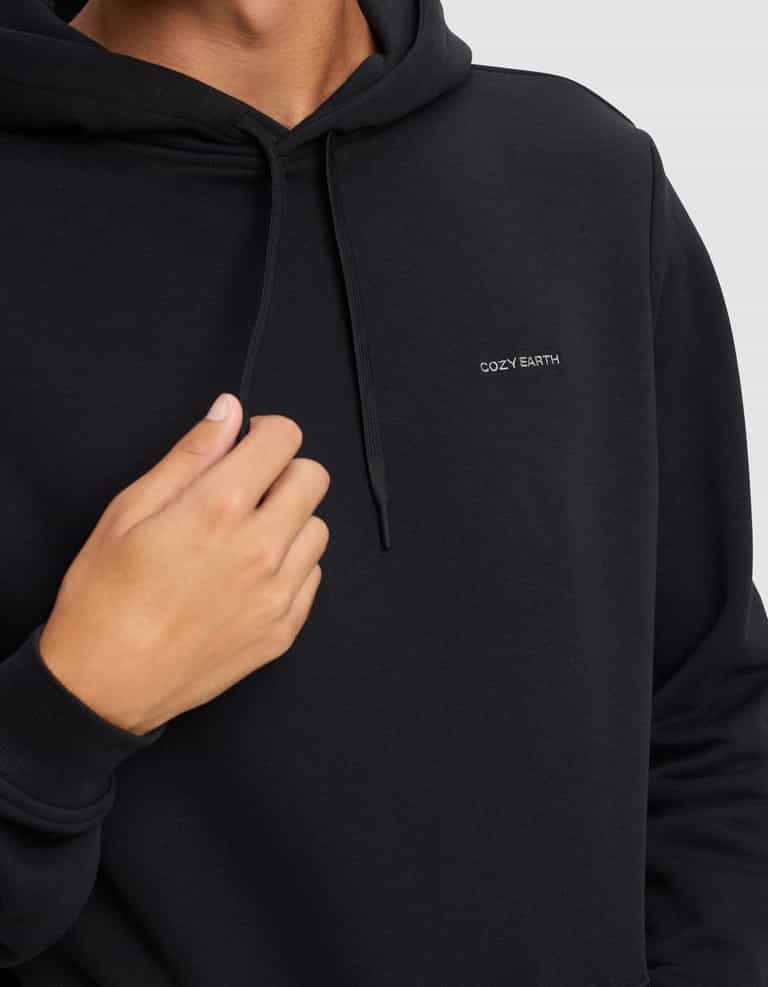 A man wears the Men's Cityscape 2.0 Hoodie by Cozy Earth, featuring "COZY EARTH" embroidered in white on the chest, holding the drawstring with one hand against a plain light background.|Color:Jet Black