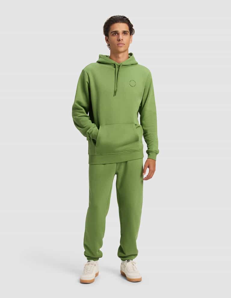 A young man wears the Cozy Earth Men's Cityscape 2.0 Hoodie & Sweatpant Set with white sneakers, standing against a plain background with his hands in the hoodie pocket and a neutral expression.|Color:Clover